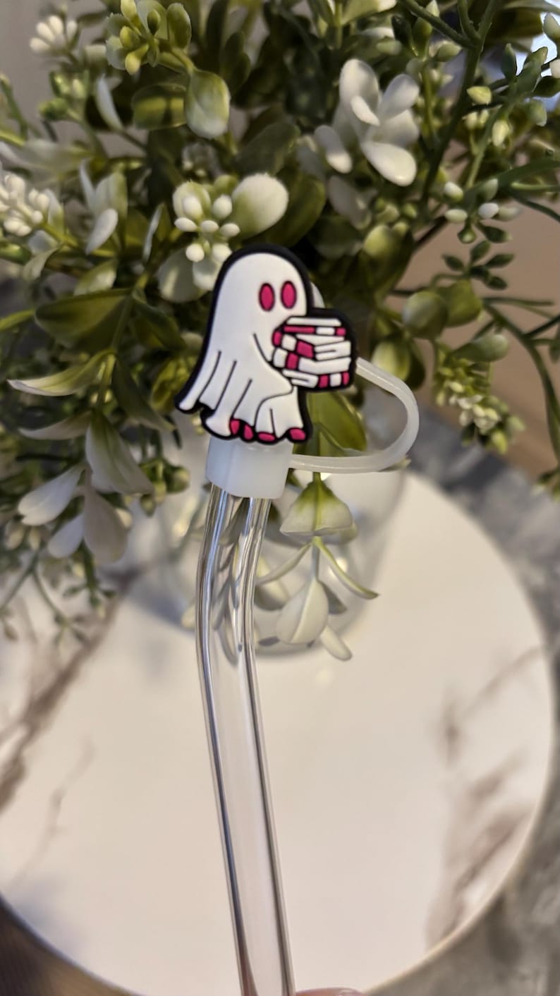 Puede incluir: Una pajita de vidrio transparente con un amuleto de fantasma blanco que sostiene una pila de libros rosas y blancos. El amuleto est&aacute; unido a la pajita con un anillo blanco.