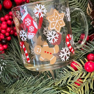 May include: A clear glass mug with a festive Christmas design. The mug features gingerbread men, candy canes, snowflakes, and a stocking. The mug is surrounded by a Christmas wreath with red berries and pine needles.