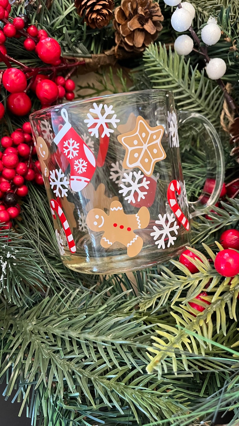 May include: A clear glass mug with a festive Christmas design. The mug features gingerbread men, candy canes, snowflakes, and a stocking. The mug is surrounded by a Christmas wreath with red berries and pine needles.