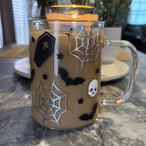 Pode incluir: Uma caneca de vidro transparente com um l&iacute;quido castanho no seu interior. A caneca tem um design de Halloween preto e prateado com morcegos, teias de aranha, estrelas e um caix&atilde;o com uma cruz.