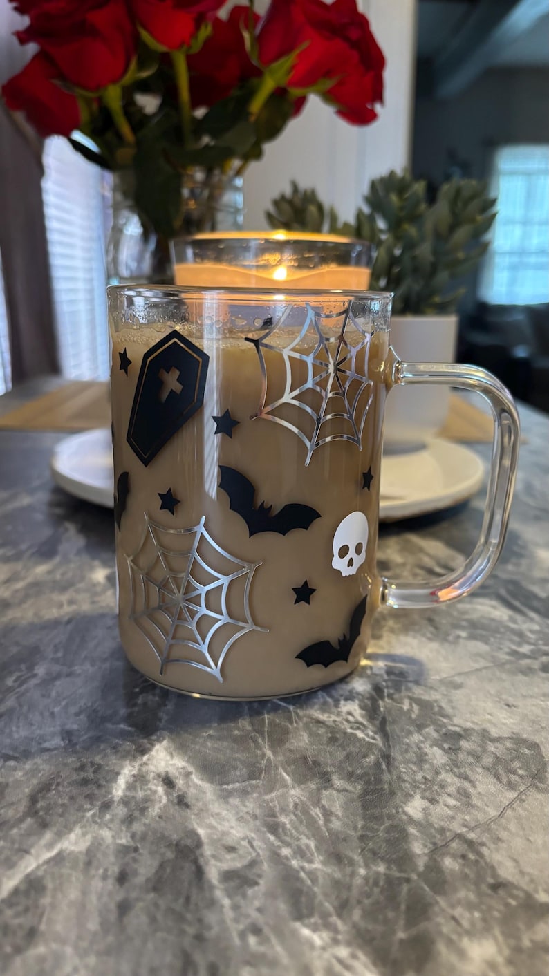 Pode incluir: Uma caneca de vidro transparente com um l&iacute;quido castanho no seu interior. A caneca tem um design de Halloween preto e prateado com morcegos, teias de aranha, estrelas e um caix&atilde;o com uma cruz.