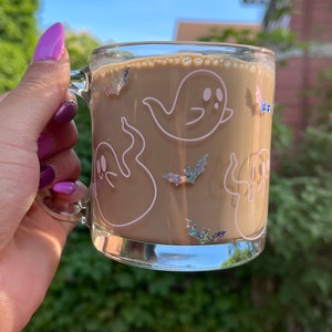 May include: A glass mug with a brown beverage inside. The mug has a white outline of ghosts and bats with iridescent glitter accents.
