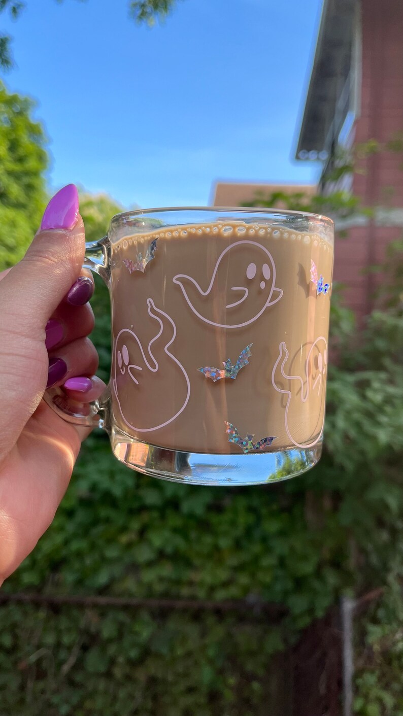 May include: A glass mug with a brown beverage inside. The mug has a white outline of ghosts and bats with iridescent glitter accents.