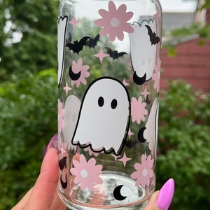 May include: A clear glass can with a pink and black ghost, flower, and crescent moon design.
