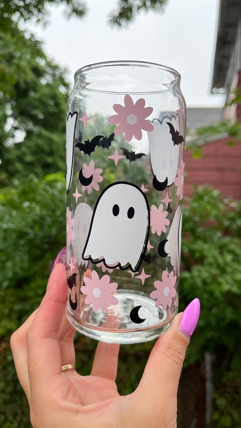 May include: A clear glass can with a pink and black ghost, flower, and crescent moon design.