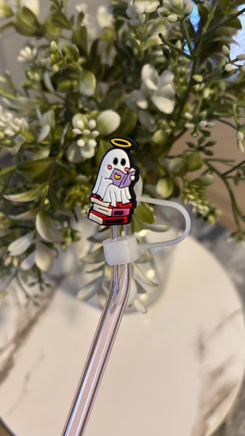 Puede incluir: Un fantasma blanco con un halo sentado en una pila de libros y leyendo un libro. El fantasma est&aacute; unido a una pajita para beber transparente con un soporte de pajita de pl&aacute;stico blanco.