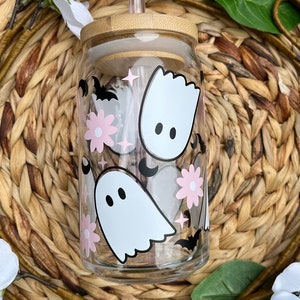 May include: A clear glass reusable water bottle with a bamboo lid and a straw. The bottle has a cute Halloween design with pink flowers, white ghosts, and black bats.