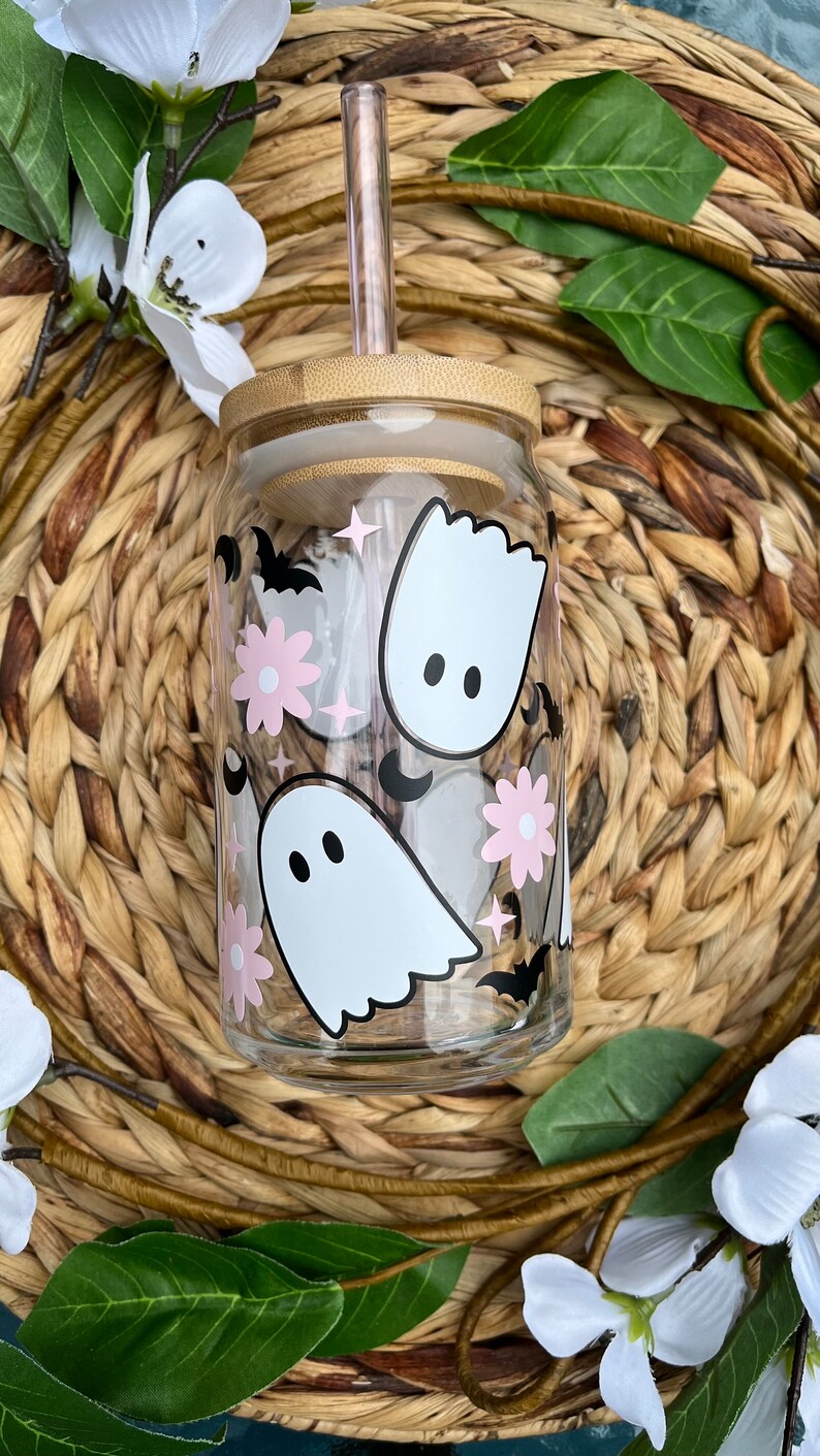 May include: A clear glass reusable water bottle with a bamboo lid and a straw. The bottle has a cute Halloween design with pink flowers, white ghosts, and black bats.