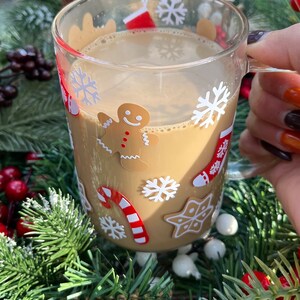 May include: A clear glass mug with a festive Christmas design featuring snowflakes, candy canes, and a gingerbread man. The mug is filled with a warm brown beverage.