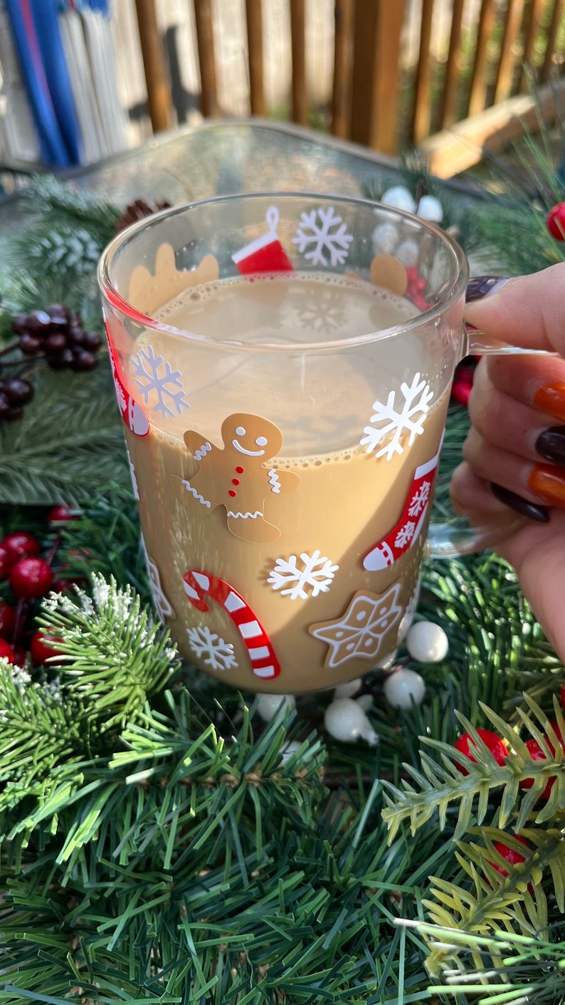 May include: A clear glass mug with a festive Christmas design featuring snowflakes, candy canes, and a gingerbread man. The mug is filled with a warm brown beverage.