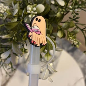 Puede incluir: Una pajita blanca con un amuleto de fantasma rosa y blanco que sostiene un libro rosa. El amuleto est&aacute; unido a la pajita con una cuerda blanca.