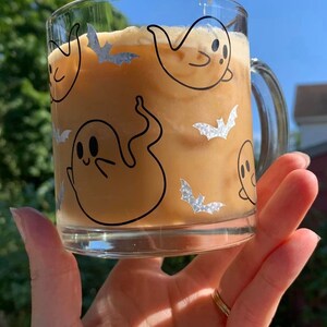 May include: A glass mug with a brown iced coffee drink. The mug has a black and white design of ghosts and bats.