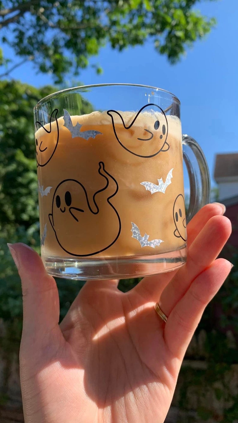 May include: A glass mug with a brown iced coffee drink. The mug has a black and white design of ghosts and bats.