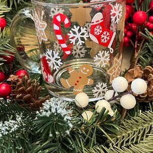 May include: A clear glass mug with a white handle features a festive Christmas design. The mug is decorated with snowflakes, candy canes, gingerbread men, and stars.