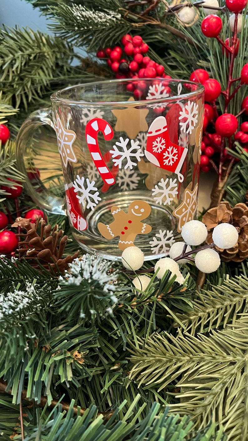 May include: A clear glass mug with a white handle features a festive Christmas design. The mug is decorated with snowflakes, candy canes, gingerbread men, and stars.