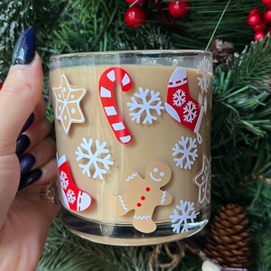 May include: A clear glass mug with a brown liquid inside. The mug is decorated with Christmas-themed stickers, including snowflakes, candy canes, gingerbread men, and stockings.