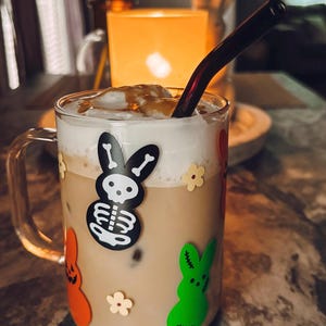 Può includere: Una tazza di vetro trasparente riempita con una bevanda marrone chiaro, sormontata da schiuma bianca e una cannuccia scura. La tazza è decorata con adesivi colorati di coniglietti e fiori. Una candela accesa è sullo sfondo.