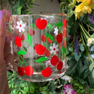 May include: A clear glass mug with a red and green cherry design. The mug has a white handle and is filled with cherries and white flowers.