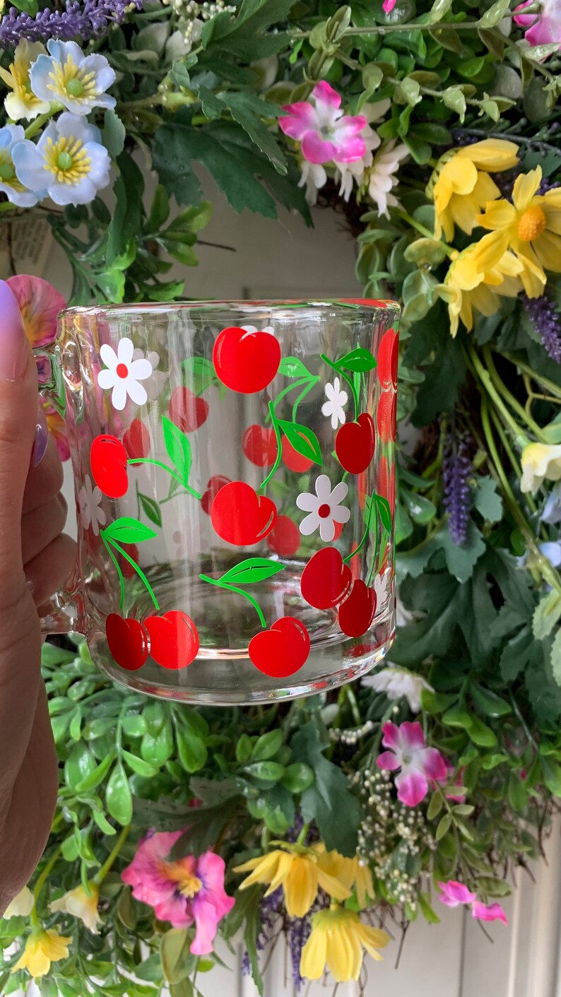 May include: A clear glass mug with a red and green cherry design. The mug has a white handle and is filled with cherries and white flowers.