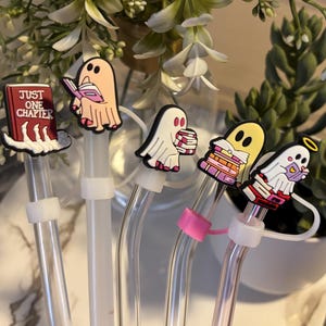 Puede incluir: Cinco pajitas de vidrio transparente con tapas de fantasmas de colores. Las tapas representan fantasmas leyendo libros, uno de ellos leyendo un libro titulado "Just One Chapter".