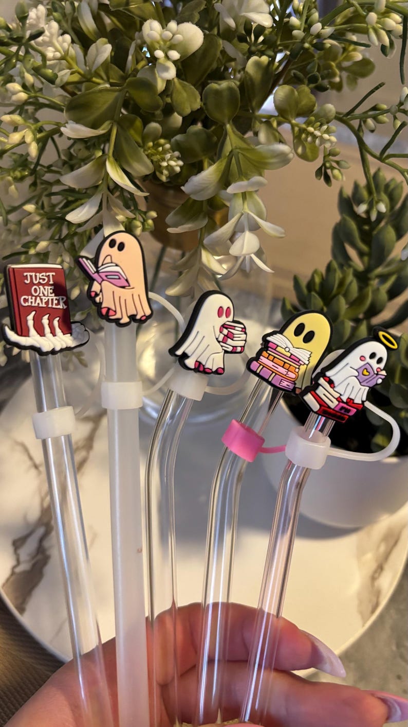 Puede incluir: Cinco pajitas de vidrio transparente con tapas de fantasmas de colores. Las tapas representan fantasmas leyendo libros, uno de ellos leyendo un libro titulado "Just One Chapter".
