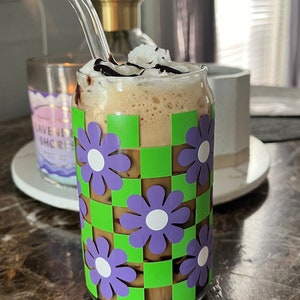 Puede incluir: Un vaso de café helado con crema batida y jarabe de chocolate en una taza a cuadros verde y morado con flores.