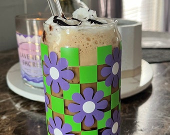 May include: A glass of iced coffee with whipped cream and chocolate syrup in a green and purple checkered cup with flowers.