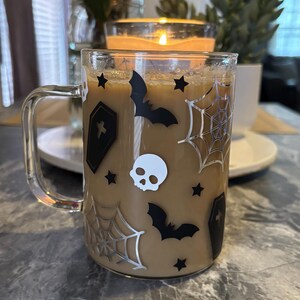 Pode incluir: Uma caneca de vidro transparente com um design de Halloween em preto e branco. A caneca est&aacute; cheia de caf&eacute; e tem uma al&ccedil;a. O design inclui morcegos, caveiras, teias de aranha e estrelas.