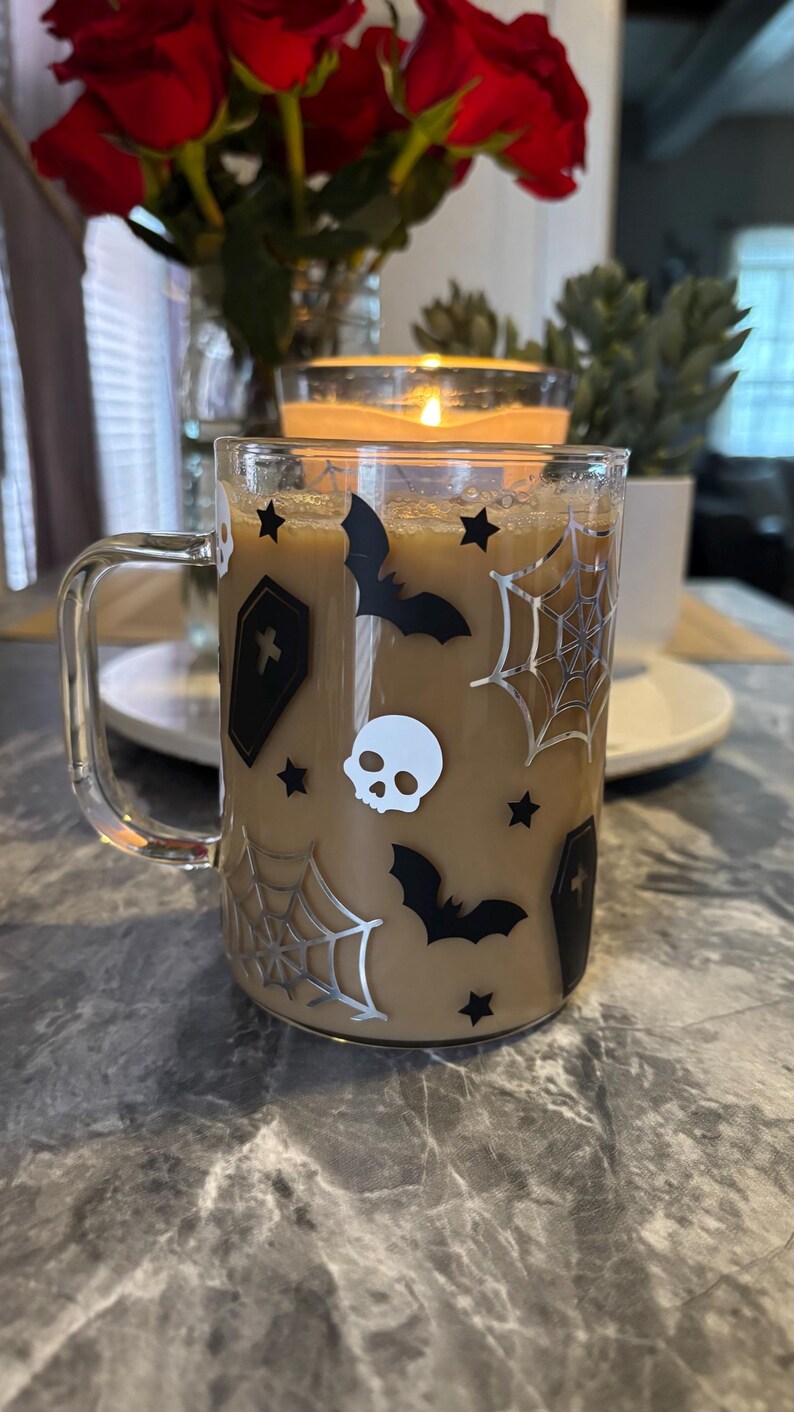 Pode incluir: Uma caneca de vidro transparente com um design de Halloween em preto e branco. A caneca est&aacute; cheia de caf&eacute; e tem uma al&ccedil;a. O design inclui morcegos, caveiras, teias de aranha e estrelas.
