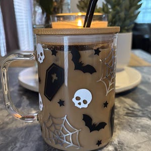Pode incluir: Uma caneca de vidro transparente com tampa de bambu e um canudo preto. A caneca tem um tema de Halloween com morcegos pretos, caveiras e teias de aranha em um fundo bege.