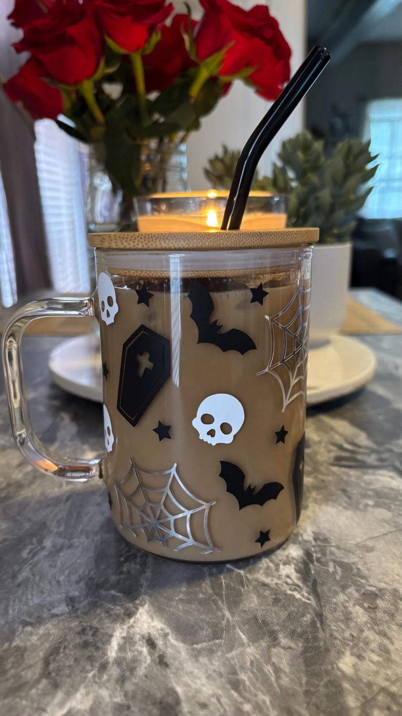 Pode incluir: Uma caneca de vidro transparente com tampa de bambu e um canudo preto. A caneca tem um tema de Halloween com morcegos pretos, caveiras e teias de aranha em um fundo bege.
