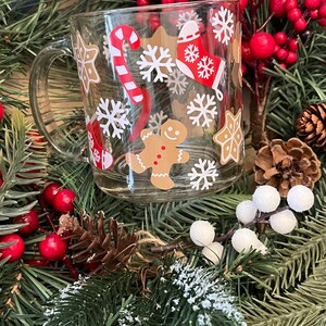 May include: A clear glass mug with a Christmas theme. The mug has a red and white candy cane, a gingerbread man, snowflakes, and a stocking with snowflakes on it.