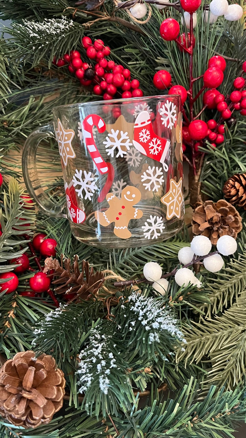 May include: A clear glass mug with a Christmas theme. The mug has a red and white candy cane, a gingerbread man, snowflakes, and a stocking with snowflakes on it.