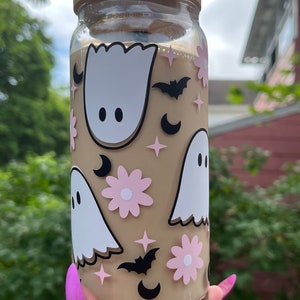 May include: A clear glass tumbler with a brown lid and a black straw. The tumbler is decorated with white ghosts, pink flowers, black bats, and black crescent moons.