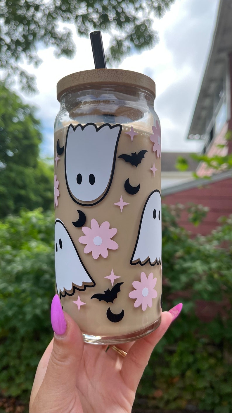 May include: A clear glass tumbler with a brown lid and a black straw. The tumbler is decorated with white ghosts, pink flowers, black bats, and black crescent moons.