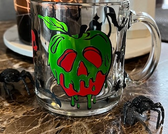 Puede incluir: Una taza de vidrio transparente con un diseño de manzana envenenada verde y roja. La manzana tiene una cara de calavera y veneno verde que gotea. La taza tiene un fondo negro con murciélagos y estrellas.