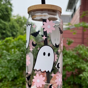 May include: A clear glass reusable water bottle with a bamboo lid and a black straw. The bottle is decorated with a pink and white ghost, flower, and crescent moon pattern.