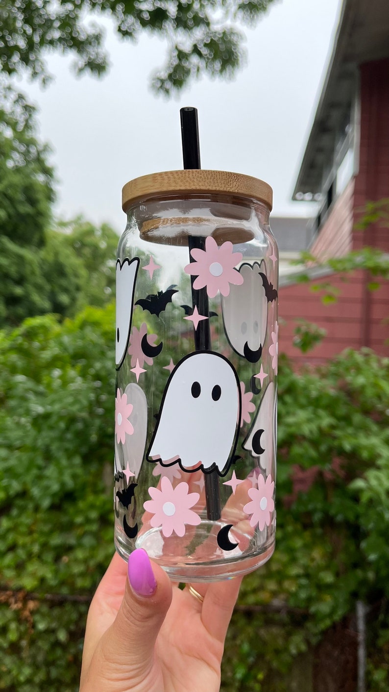 May include: A clear glass reusable water bottle with a bamboo lid and a black straw. The bottle is decorated with a pink and white ghost, flower, and crescent moon pattern.