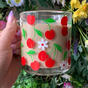 May include: A glass mug with a white background and red cherries and white flowers printed on it. The mug is filled with a light brown beverage.