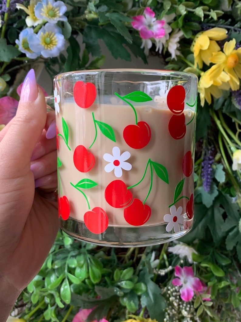 May include: A glass mug with a white background and red cherries and white flowers printed on it. The mug is filled with a light brown beverage.