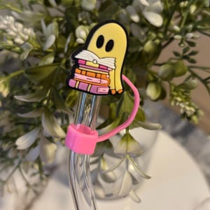 Puede incluir: Una pajita de vidrio transparente con un soporte de pajita de silicona rosa y un amuleto de fantasma amarillo que sostiene una pila de libros rosas y naranjas.