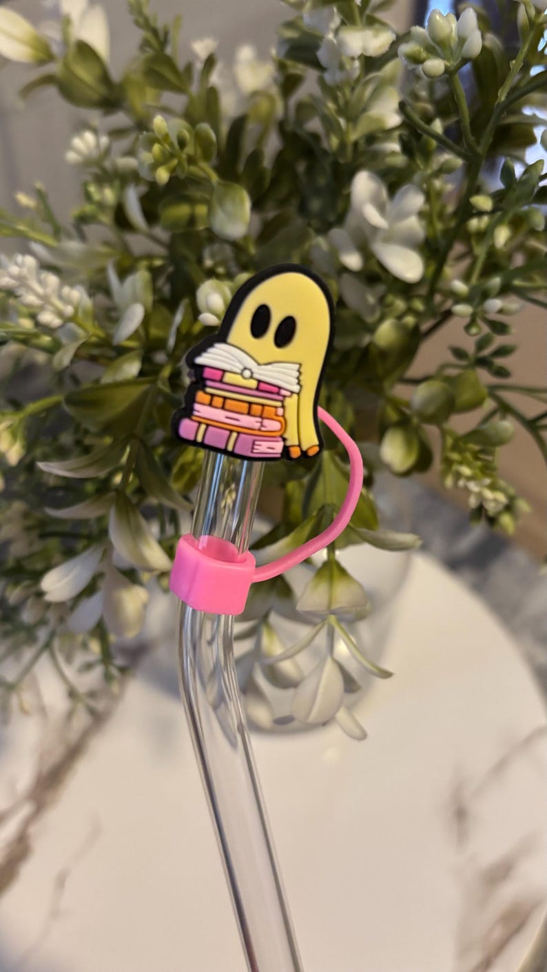 Puede incluir: Una pajita de vidrio transparente con un soporte de pajita de silicona rosa y un amuleto de fantasma amarillo que sostiene una pila de libros rosas y naranjas.