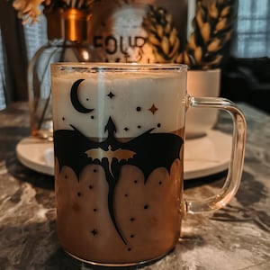 Op de afbeelding: Een glazen mok met een ontwerp van een zwarte draak en gouden sterren. De mok is gevuld met een lichtbruine drank met een witte schuimlaag.