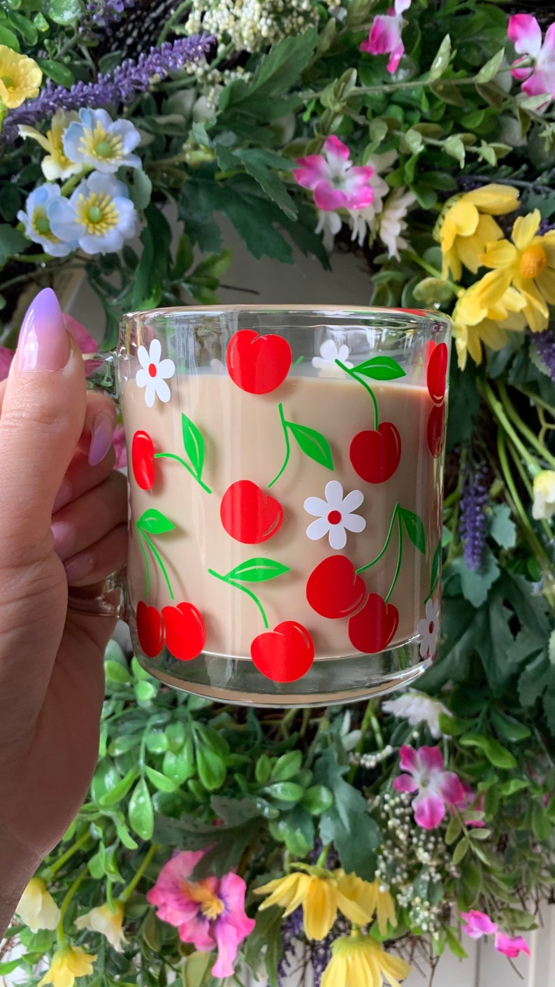 May include: A glass mug with a cherry and flower design. The mug is filled with a light brown beverage.