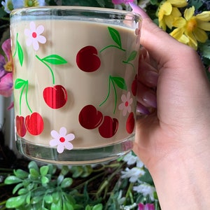 May include: A glass mug with a beige background and a pattern of red cherries and white flowers. The mug is filled with a light brown liquid.