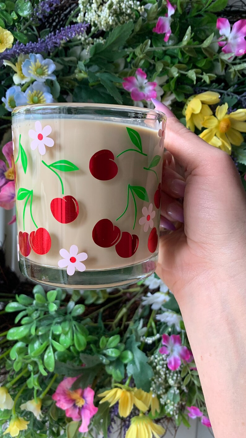 May include: A glass mug with a beige background and a pattern of red cherries and white flowers. The mug is filled with a light brown liquid.
