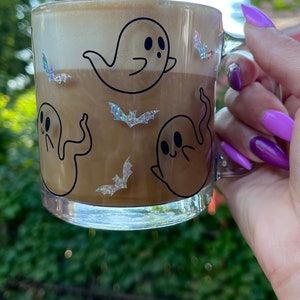 May include: A clear glass mug with a brown liquid and a white foam top. The mug has a black and white design of ghosts and bats. The mug is being held by a hand with purple nail polish.