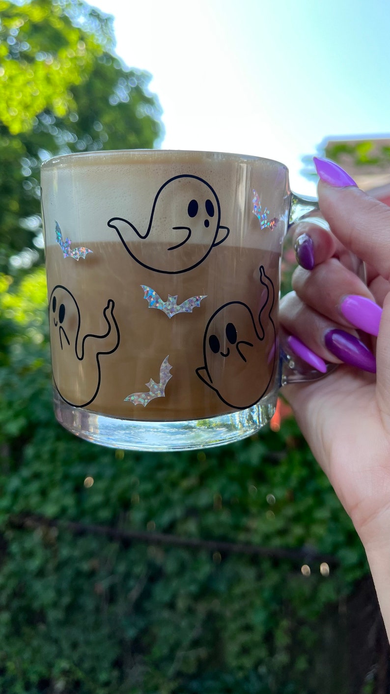 May include: A clear glass mug with a brown liquid and a white foam top. The mug has a black and white design of ghosts and bats. The mug is being held by a hand with purple nail polish.