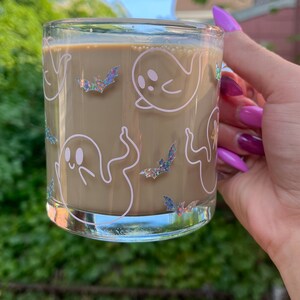 May include: A glass mug with a brown beverage inside. The mug has a white design of ghosts and bats with iridescent glitter accents.
