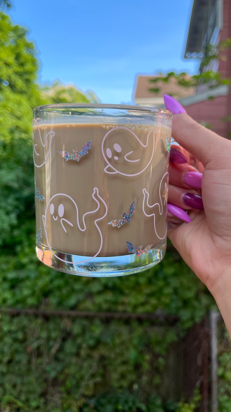 May include: A glass mug with a brown beverage inside. The mug has a white design of ghosts and bats with iridescent glitter accents.